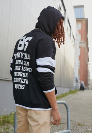 Młoda osoba z czerwonymi dredami, ubrana w czarną bluzę z białymi paskami i beżowe spodnie, stojąca przy barierce na miejskiej ulicy.