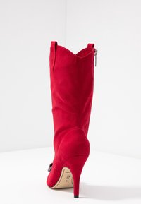 Botte haute en similicuir suédé rouge à talon aiguille avec un bout pointu, un haut festonné et une fermeture éclair sur le côté ; présente un talon stiletto et une finition texturée.