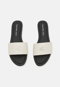 Sandales à glisser Calvin Klein avec une large sangle blanc cassé, semelle noire et détail de logo en relief. Matériau doux avec un design plat.