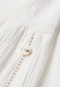 Tela blanca texturizada con pliegues verticales, que presenta una insignia redonda en tono dorado con el logo de "Superdry". Detalle en primer plano.