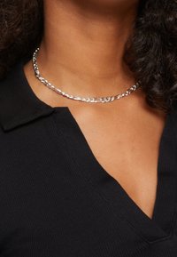 Silberne Kettenhalskette mit länglichen Gliedern, getragen von einer Person in einem schwarzen Kragenoberteil. Glatte Textur, polierte Oberfläche.