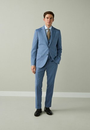 Bărbat îmbrăcat într-un costum albastru în trei piese, cu cravată aurie și pantofi negri, pe un fundal simplu de culoare gri deschis.