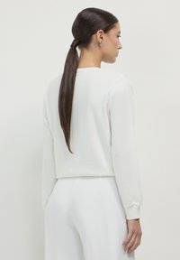 Frau mit einem langen braunen Pferdeschwanz, die ein weißes Langarmoberteil und weiße Hosen trägt, seitlich gegen einen schlichten weißen Hintergrund blickend.