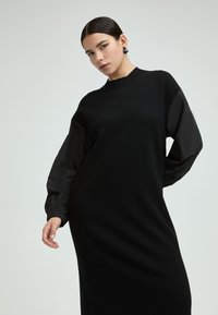 Schwarzes Kleid mit figurbetonter Silhouette, langen schwarzen Stoffärmeln und strukturierten Bündchen. Glattes Material mit hohem Kragen.