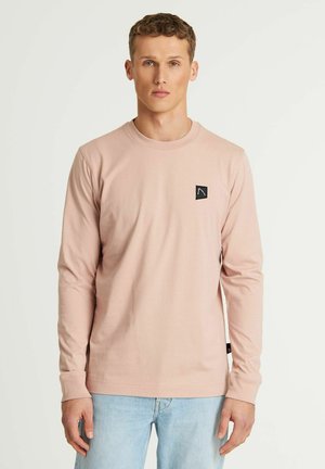 Lyserød langærmet t-shirt lavet af blød bomuld, med et lille sort logo-patch på venstre bryst og en rund halsudskæring.