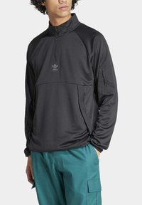 Svart zip-up pullover med hög krage och textilstruktur. Har en logotyp och en zippad ficka på vänster ärm.