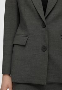 Blazer gris avec deux boutons noirs, design à une seule rangée de boutons, revers crantés, et un tissu texturé comportant de petits motifs subtils.