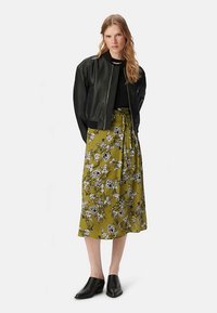 Czarna skórzana kurtka typu bomber na czarnej bluzce, zestawiona z oliwkowozieloną spódnicą w kwiaty do kolan i czarnymi klapkami.