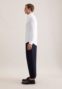 Camicia bianca a maniche lunghe, pantaloni blu navy, scarpe nere. La camicia presenta un colletto a punta e sottili spacchi laterali, tessuto liscio e vestibilità comoda.