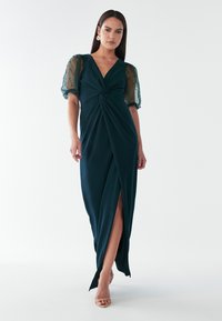 Garšīgs, smaragda zaļš kleitas modelis ar dziļu V-veida dekoltē un savērpendi, kam ir caurspīdīgas puff piedurknes ar pogām un augstu šķēlumu līdz augšstilbam.