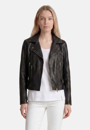 Blonde Frau mit glatten Haaren, die eine schwarze Lederjacke über einem weißen Hemd und weißen Hosen trägt, vor einem einfarbigen Hintergrund stehend.