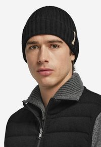 Czarna prążkowana dzianinowa czapka beanie z teksturowanym wykończeniem i małym logo; noszona z czarno-szarą kamizelką na zamek błyskawiczny z wysokim kołnierzem.