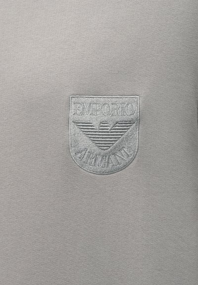 Tecido cinza de malha apresenta um logótipo texturado bordado com a inscrição "Emporio Armani" em fio prateado no peito, com elementos de design de asas e riscas.