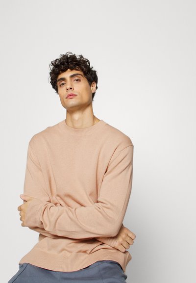 Suéter de malha rosa claro com mangas longas, punhos e barra em canelado. Modelo sentado, com os braços cruzados, cabelo curto e encaracolado, em fundo simples.