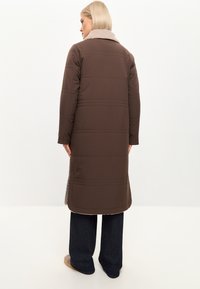 Brun pufferjakke med bælte-design, der har en blød beige krave. Lange ærmer, knælang og med sideskår for bedre bevægelsesfrihed.