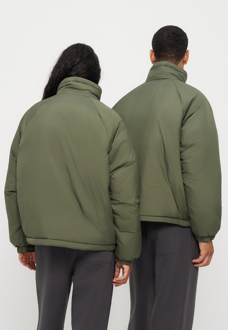Δύο άτομα από πίσω, που φορούν πράσινα επενδυμένα μπουφάν και σκούρα παντελόνια απέναντι σε ένα απλό ανοιχτό φόντο.