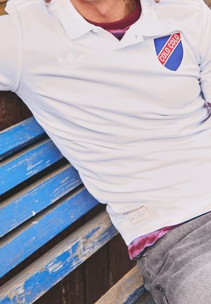 Person iført en hvid Colo-Colo polo med et rødt og blåt mærke, siddende på en vejrbidt blå træbænk.