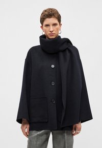Tmavě modrý kabát ze směsi vlny s vysokým límcem, zapínáním na knoflíky vpředu a bočními kapsami. Má nadměrný, kokonový tvar a jemnou texturu.