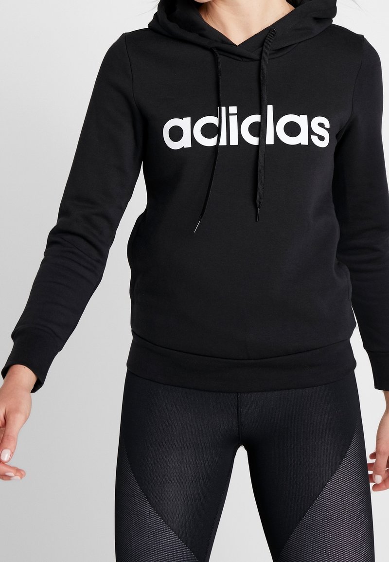 Persona che indossa un hoodie nero Adidas con logo bianco e leggings neri texture, in piedi con le braccia leggermente estese.