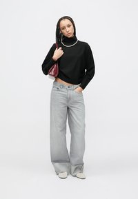 Černý crop top s dlouhým rukávem, světlé šedé široké džíny s vysokým pasem, červená kabelka a světlé boty s hladkou texturou, postava stojící na jednobarevném pozadí.