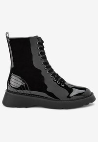Botín negro de charol brilloso con suela de goma gruesa, cordones en la parte frontal con ojales de metal y tirador en la parte trasera.