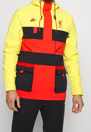 Homme portant une veste Nike jaune, rouge et noire avec le logo du Liverpool FC et plusieurs poches frontales, assortie à un pantalon noir.