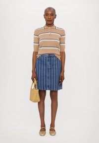 Polo shirt rayé beige, jupe en denim à rayures verticales, sac tissé jaune, chaussures plates dorées, mannequin posé contre un fond neutre.