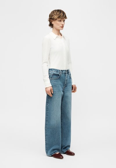 Γυναίκα που φοράει εφαρμοστό λευκό πουκάμισο με κουμπιά, φαρδιά μπλε τζιν παντελόνια και καφέ μυτερά παπούτσια, στέκεται μπροστά σε απλό ανοιχτό φόντο.