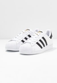 Baskets blanches avec un embout texturé, trois bandes noires et un onglet de talon noir. Comprend des lacets blancs et un logo doré sur la languette.