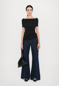 Zwarte off-shoulder top gecombineerd met wijde, donkere denim broeken met een voorzijde-split; model houdt een zwarte handtas vast.