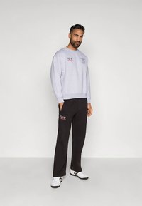 Homem de pé contra um fundo simples, vestindo um suéter cinza claro e calças pretas com tênis brancos, com as mãos nos bolsos, olhando em frente.