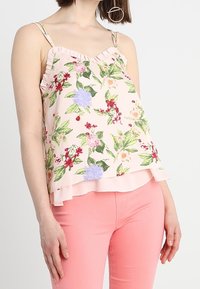 Blusa floral rosa con bordes rizados, que presenta diversas flores coloridas y hojas verdes sobre un fondo claro. Combinada con pantalones rosas.