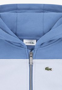 Φούτερ με φερμουάρ και δίχρωμο σχέδιο σε ανοιχτό και σκούρο μπλε, κατασκευασμένο από βαμβάκι. Διαθέτει μικρό λογότυπο Lacoste και ασημένια εξαρτήματα φερμουάρ.