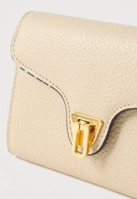 Beżowy portfel ze skóry teksturowanej z zakrzywioną klapką i błyszczącym złotym trójkątnym zapięciem na neutralnym tle.