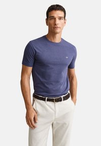 Mand iført en tætsiddende navyblå t-shirt med korte ærmer og lille logo på brystet, hvide bukser og brun flettet bælte, stående med hænderne i lommerne.