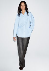 Lichtblauw oversized overhemd met lange mouwen, gecombineerd met donkergrijze wijde broekspijpen, gedragen met open zwarte sandalen.