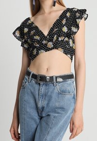 Top court noir avec imprimé marguerite et pois blancs, manches à volants, associé à un jean taille haute bleu clair et une ceinture noire.