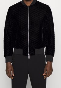 Svart kviltad bomberjacka i mjuk sammet med ribbade muddar och nederkant, med dragkedja fram och sidofickor.