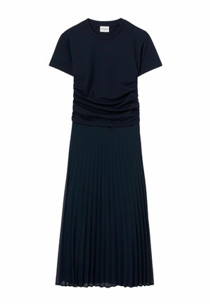 Donna con top blu navy a maniche corte e gonna midi plissettata con sandali dorati con tacco a blocchetto, in piedi davanti a uno sfondo semplice.
