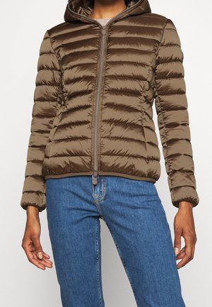 Person iført en brun dunjakke med vandrette syninger og blå jeans, stående mod en ensfarvet lys baggrund.