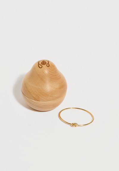 Ξύλινο δοχείο σε σχήμα αχλαδιού με καπάκι δίπλα σε λεπτό χρυσό βραχιόλι με μικρή λεπτομέρεια κόμπου σε λευκό φόντο.