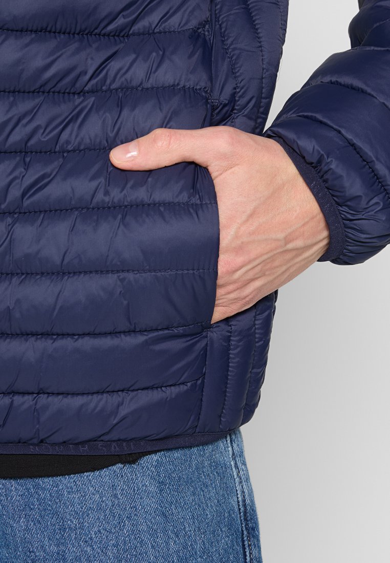 Ręka włożona w boczną kieszeń granatowej pikowanej kurtki, w zestawieniu z niebieskimi dżinsami.