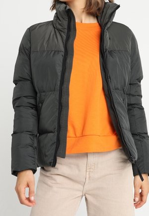 Personne portant une doudoune noire ouverte sur un sweat-shirt orange et un pantalon beige sur un fond uni.