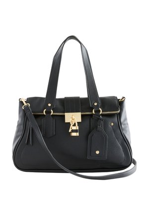 Frau trägt graues kurzärmeliges Oberteil und braune Hose, hält schwarze Handtasche mit goldenem Schloss und trägt goldene Creolen und klobige Armbänder.