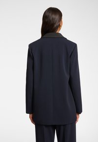 Blazer navy con cuello negro, corte entallado, mangas largas y una textura suave, mostrado desde la parte de atrás con un mínimo de detalles visibles.