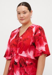 Vestido floral vermelho com mangas curtas e ligeiras e um decote em V profundo. O tecido apresenta grandes rosas misturadas em tons de vermelho e rosa, com um detalhe de nó na cintura.
