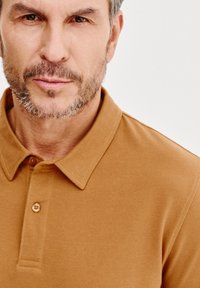 Brązowa koszulka polo z klasycznym kołnierzykiem, trzema guzikami z przodu i teksturowanym materiałem. Skupienie na górnej części klatki piersiowej i okolicy dekoltu.