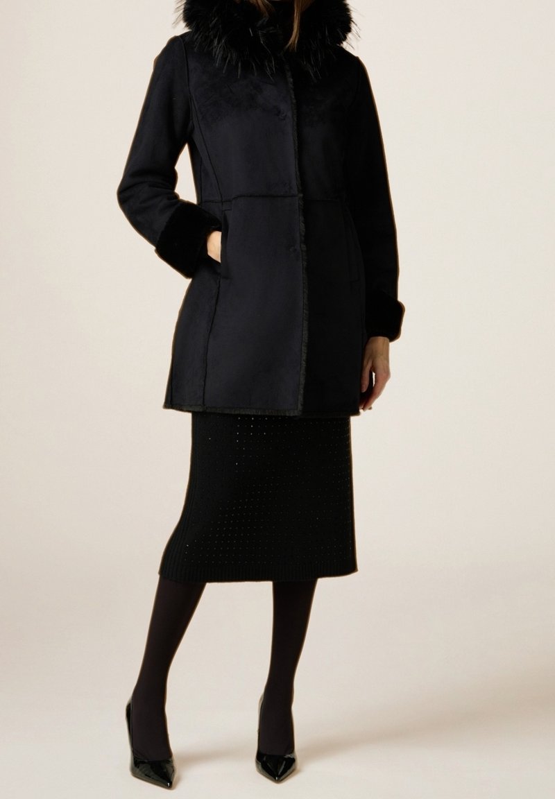 Femme portant un manteau noir mi-longueur avec col et poignets en fourrure, jupe noire, collants et talons aiguilles, debout sur un fond neutre.