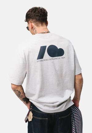 Mężczyzna z tatuażami i łańcuszkiem na szyi nosi szarą koszulkę z ciemnoniebieską grafiką i napisem „Established in Poland in 1994” na plecach.