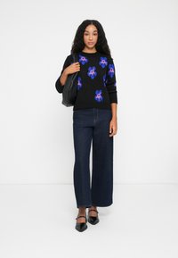 Zwarte trui met blauwe en roze bloemenpatronen, gecombineerd met een wijde, donkere spijkerbroek. Accessoires omvatten een zwarte tote bag en puntige flats.